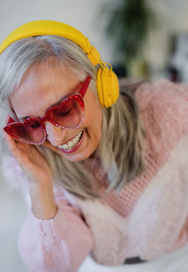 Portrait of cheerful senior woman with sunglasses and headphones indoors at home, laughing.