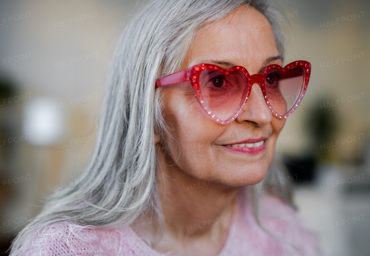 A portrait of happy senior woman with party sunglasess standing indoors at home.