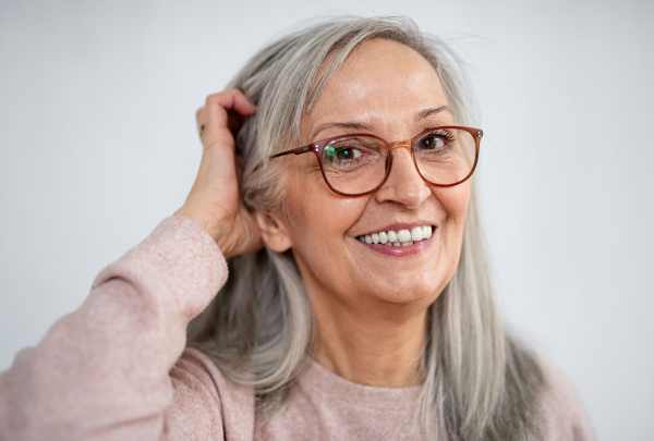 Front view of portrait of senior woman standing indoors against light background, looking at camera.