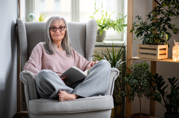 Portrait of senior woman sitting indoors on sofa at home, relaxing and reading book.