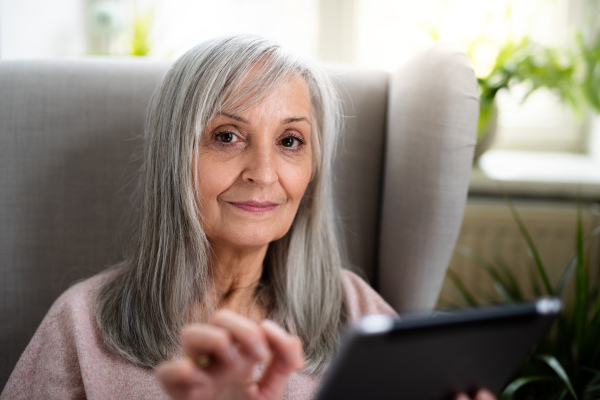 Portrait of happy senior woman sitting indoors on sofa at home, using tablet.