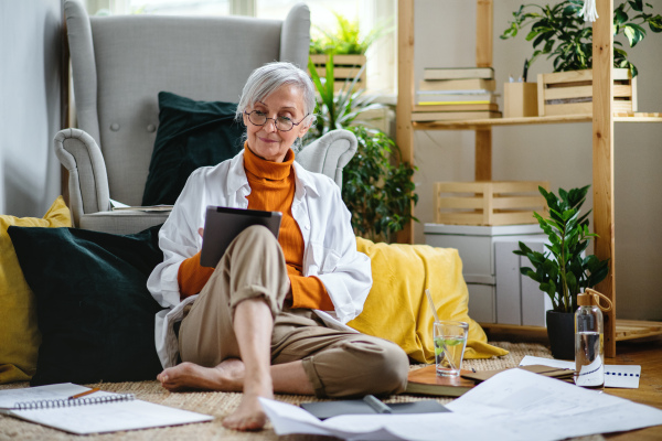 Portrait of senior woman architect sitting with blueprints on floor indoors at home, working.