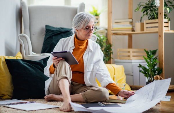 Portrait of senior woman architect sitting with blueprints on floor indoors at home, working.