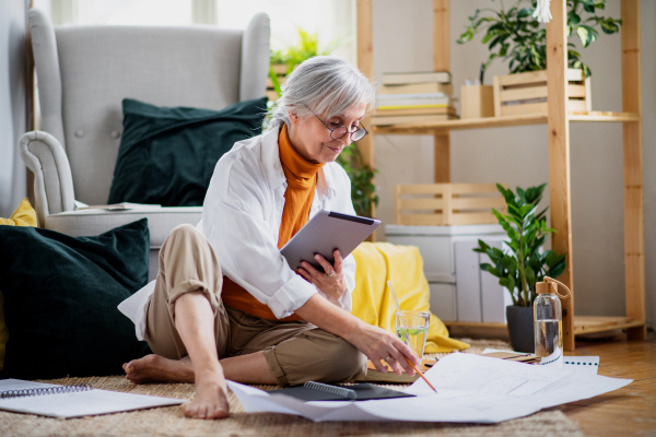 Portrait of senior woman architect sitting with blueprints on floor indoors at home, working.