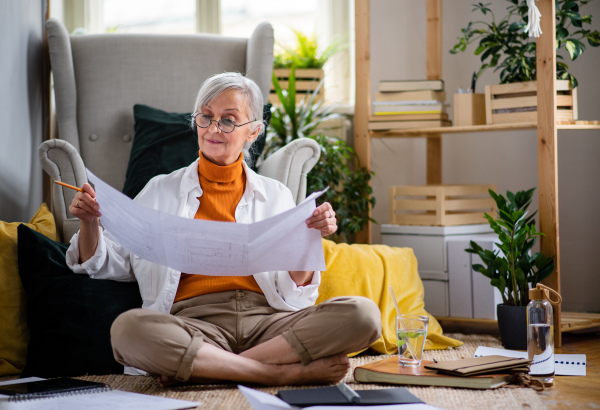 Portrait of senior woman architect sitting with blueprints on floor indoors at home, working.