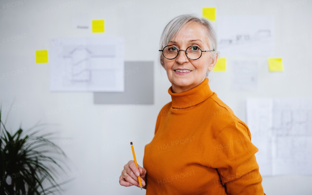 Portrait of senior woman architect with blueprints standing indoors at home, working.