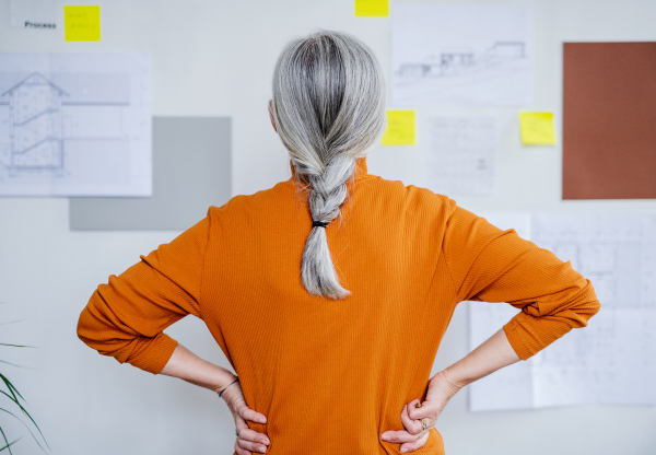 A rear view of senior woman architect standing indoors at home, standing and working.