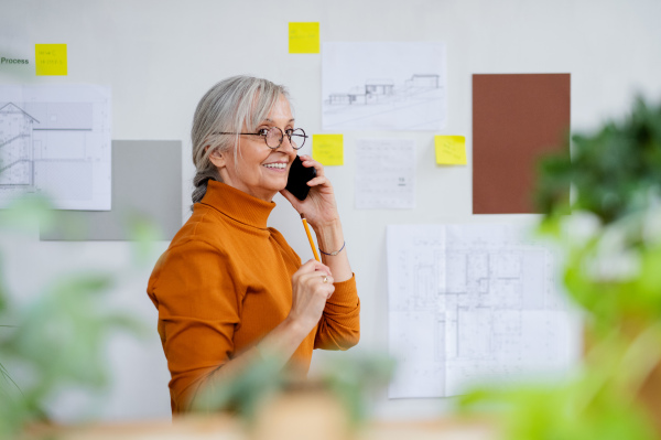 Portrait of senior woman architect with blueprints and smartphone standing indoors at home, working.