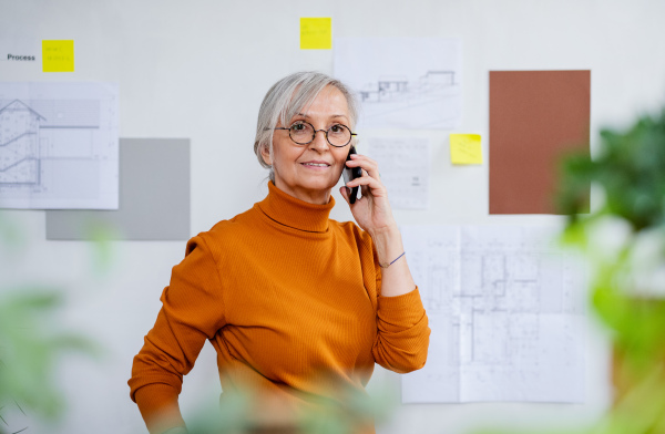 A rear view of senior woman architect standing indoors at home, using telephone.