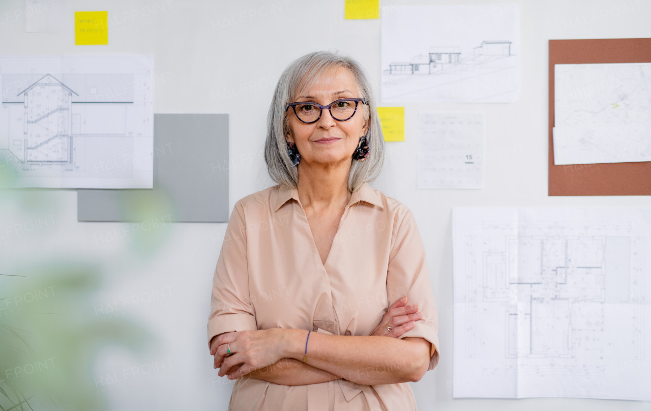 Portrait of senior woman architect with blueprints standing indoors at home, working.