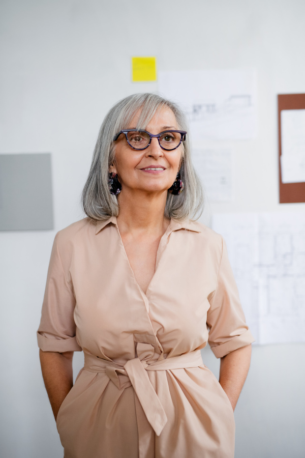 Front view of beautiful senior woman architect standing indoors at home, hands in pockets.