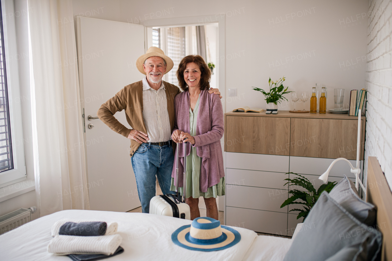 A happy senior couple with suitcase standing in hotel room, looking at camera