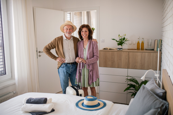 A happy senior couple with suitcase standing in hotel room, looking at camera