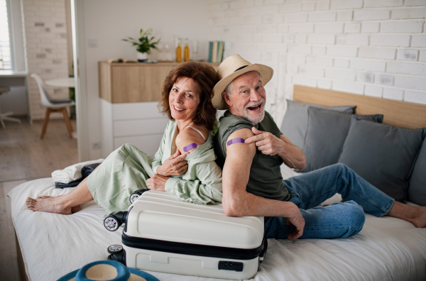 Happy senior couple sitting on bed indoors in hotel room, coronavirus vaccination, holiday and new normal concept.