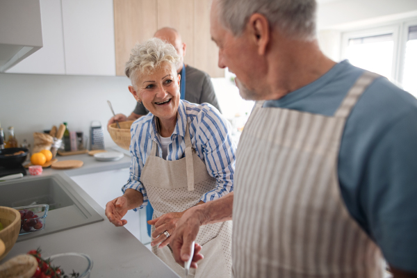 A senior couple with friends having party indoors, talking when cooking.