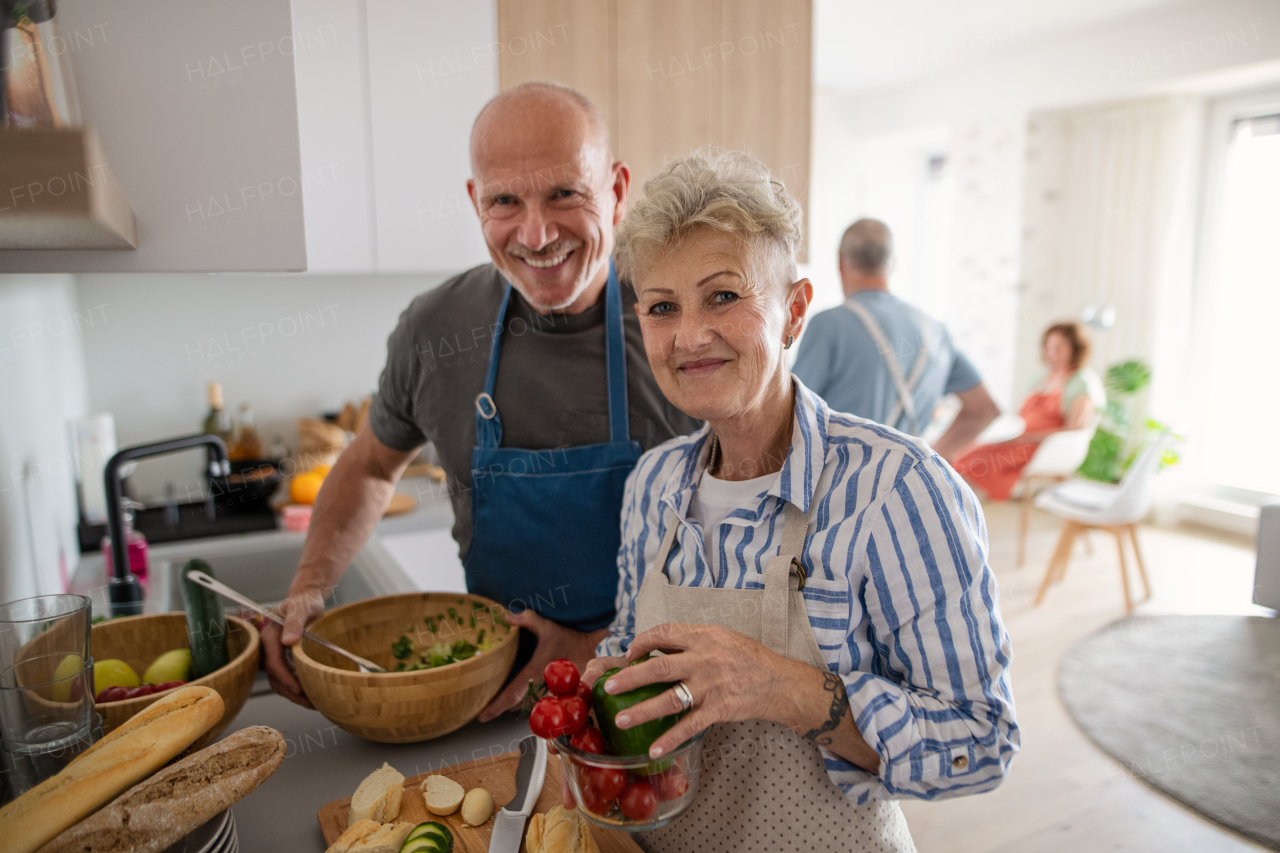 A senior couple with friends having party indoors, looking at camera when cooking.