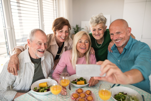 A group of senior friends having party indoors, taking selfie when eating at the table.