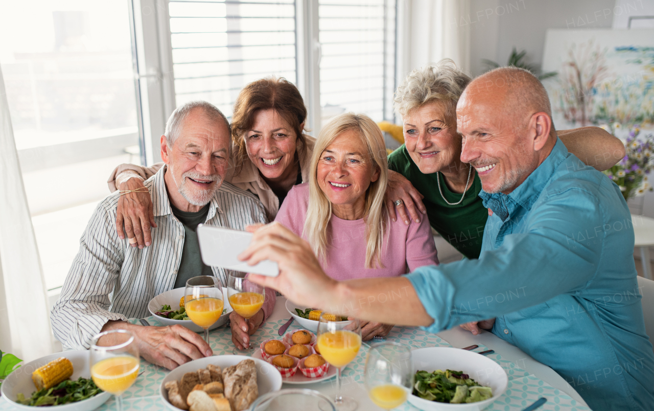 A group of senior friends having party indoors, taking selfie when eating at the table.