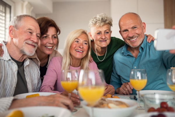 A group of senior friends having party indoors, taking selfie when eating at the table.