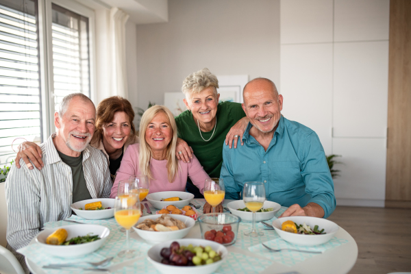 Group of cheerful senior friends having party indoors, looking at camera when eating.