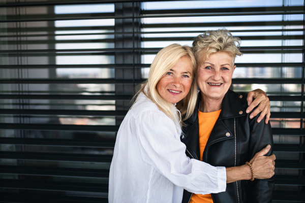 Two happy senior women friends standing outdoors in front of window, looking at camera.