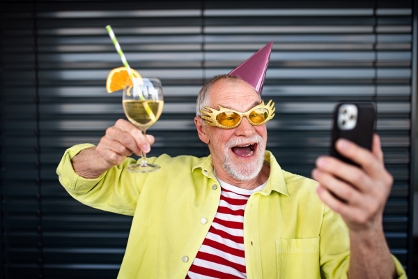 Portrait of crazy senior man with party hat and drink standing outdoors, taking selfie.