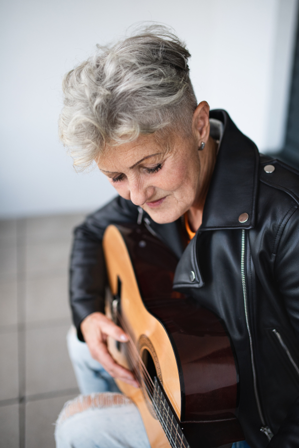 A portrait of senior woman sitting against black backround, playing guitar.