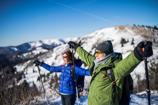 Side view of senior couple hikers with nordic walking poles in snow-covered winter nature, stretching arms.