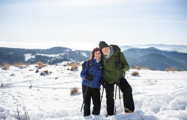 Senior couple hikers with nordic walking poles in snow-covered winter nature, looking at camera.