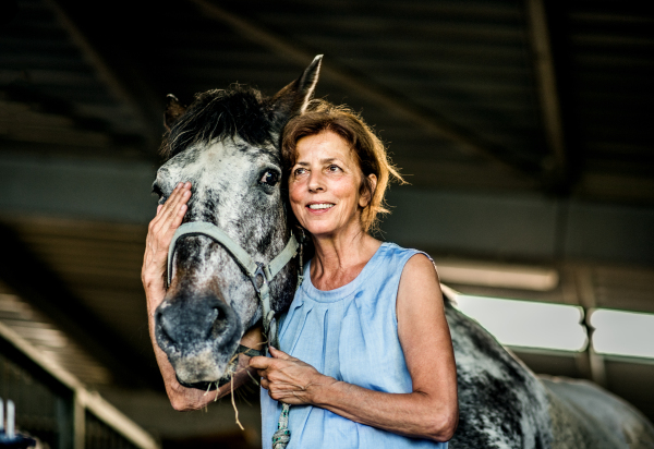 A happy senior woman with closed eyes standing close to a horse in a stable, holding it.