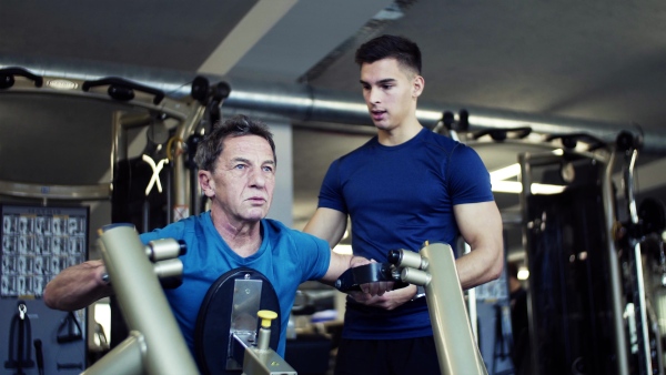 A senior man with a young male trainer doing strength workout exercise in gym.