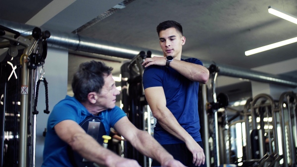 A senior man with a young male trainer doing strength workout exercise in gym.