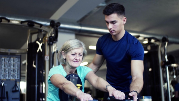 A cheerful female senior with a young trainer doing strength workout exercise in gym.