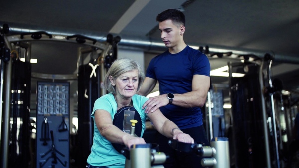 A female senior with a young trainer doing strength workout exercise in gym. Slow motion.