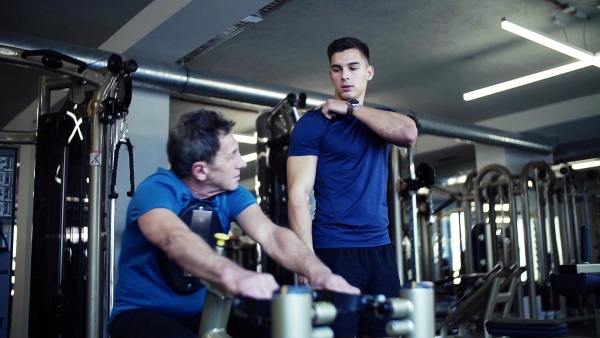 A senior man with a young male trainer doing strength workout exercise in gym. Slow motion.