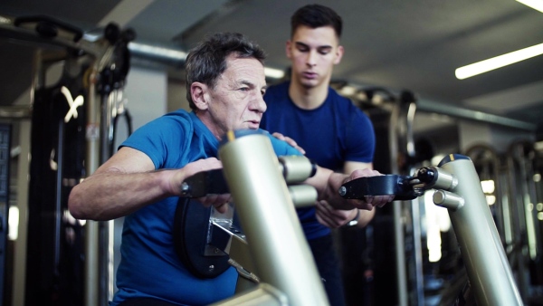 A senior man with a young male trainer doing strength workout exercise in gym. Slow motion.