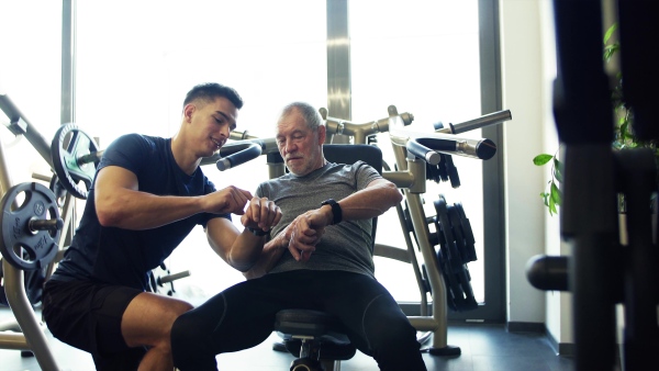 A senior man and a young male trainer in gym setting smartwatch before doing exercise.