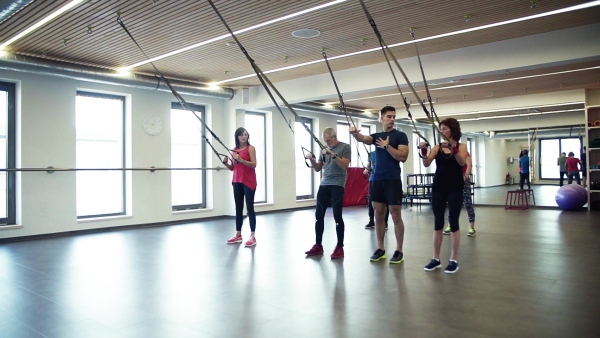 A group of cheerful seniors in gym with a young personal trainer doing exercise with TRX. Slow motion.