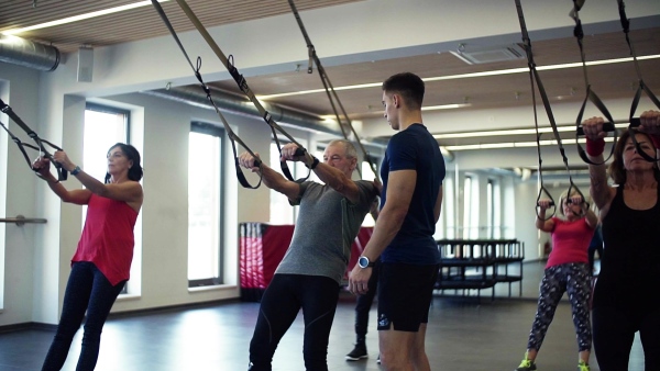 A group of cheerful seniors in gym with a young personal trainer doing exercise with TRX. Slow motion.