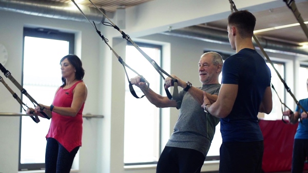 A group of cheerful seniors in gym with a young personal trainer doing exercise with TRX. Slow motion.