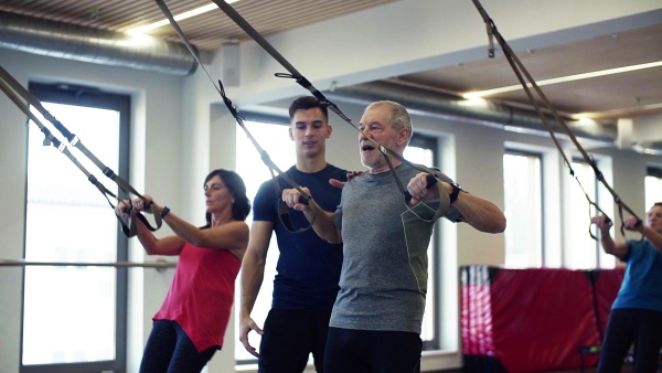 A group of cheerful seniors in gym with a young personal trainer doing exercise with TRX. Slow motion.