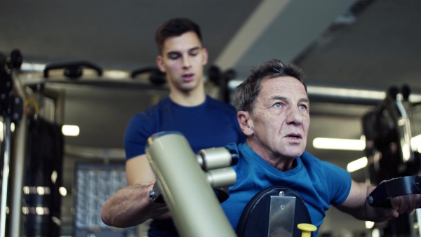 A senior man with a young male trainer doing strength workout exercise in gym.