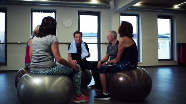 A group of cheerful seniors in gym resting after doing exercise on fit balls, talking. Slow motion.