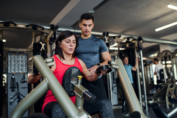 A cheerful female senior with a young trainer doing strength workout exercise in gym.