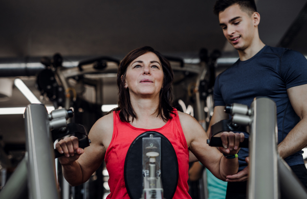 A cheerful female senior with a young trainer doing strength workout exercise in gym.