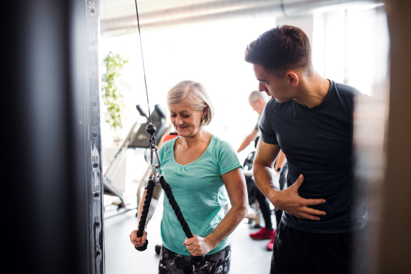 A cheerful female senior with a young trainer doing strength workout exercise in gym.