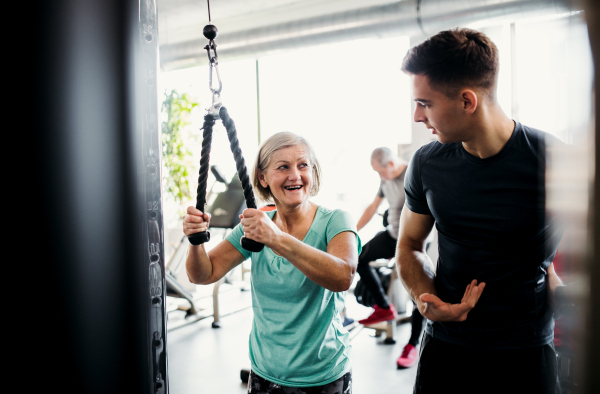 A cheerful female senior with a young trainer doing strength workout exercise in gym.