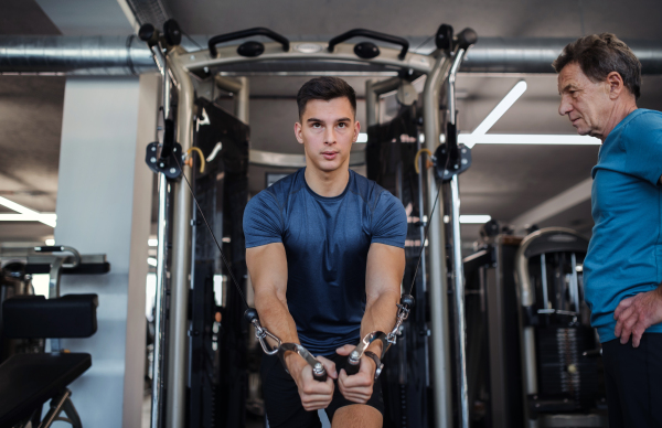 A senior man with a young male trainer doing strength workout exercise in gym.