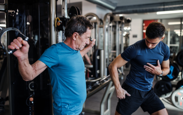 A senior man with a young male trainer doing strength workout exercise in gym.