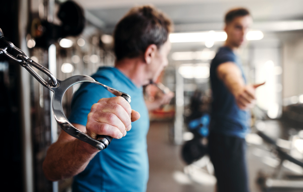 A senior man with an unrecognizable young male trainer doing strength workout exercise in gym.